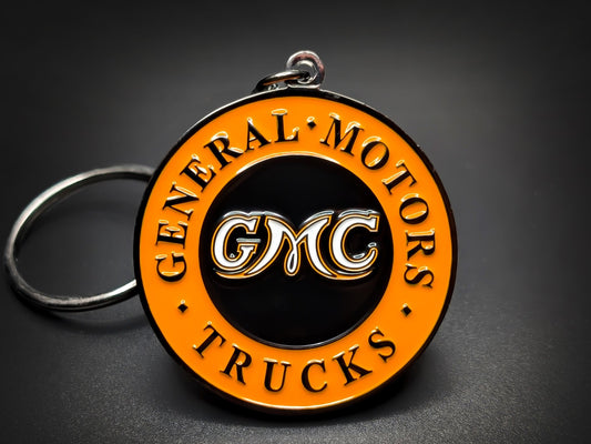 Round black and orange GMC metal double-sided keychain with "GENERAL MOTORS TRUCKS" and the classic GMC logo in white.