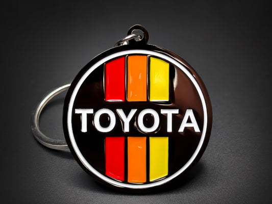 Round black metal Toyota double-sided keychain with a white logo and red, orange, and yellow vertical retro stripes.