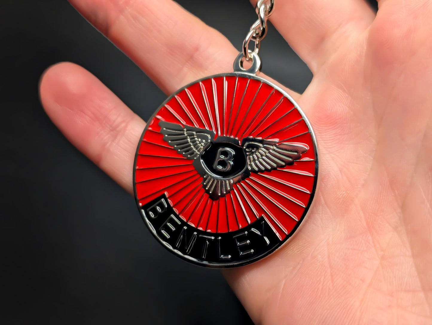 A hand holding the Bentley badge keychain, highlighting the polished metal finish and the detailed "Bentley" lettering at the bottom.