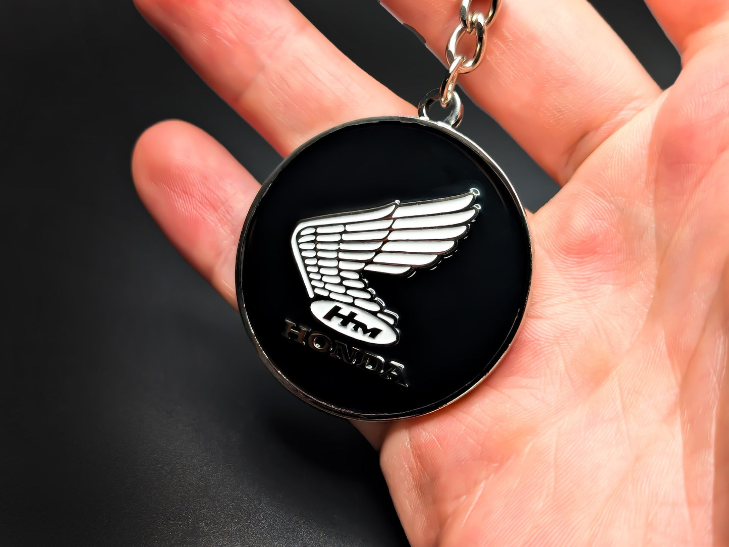 Hand holding a circular Honda heritage wing badge keychain to demonstrate the size, weight, and premium metallic finish.