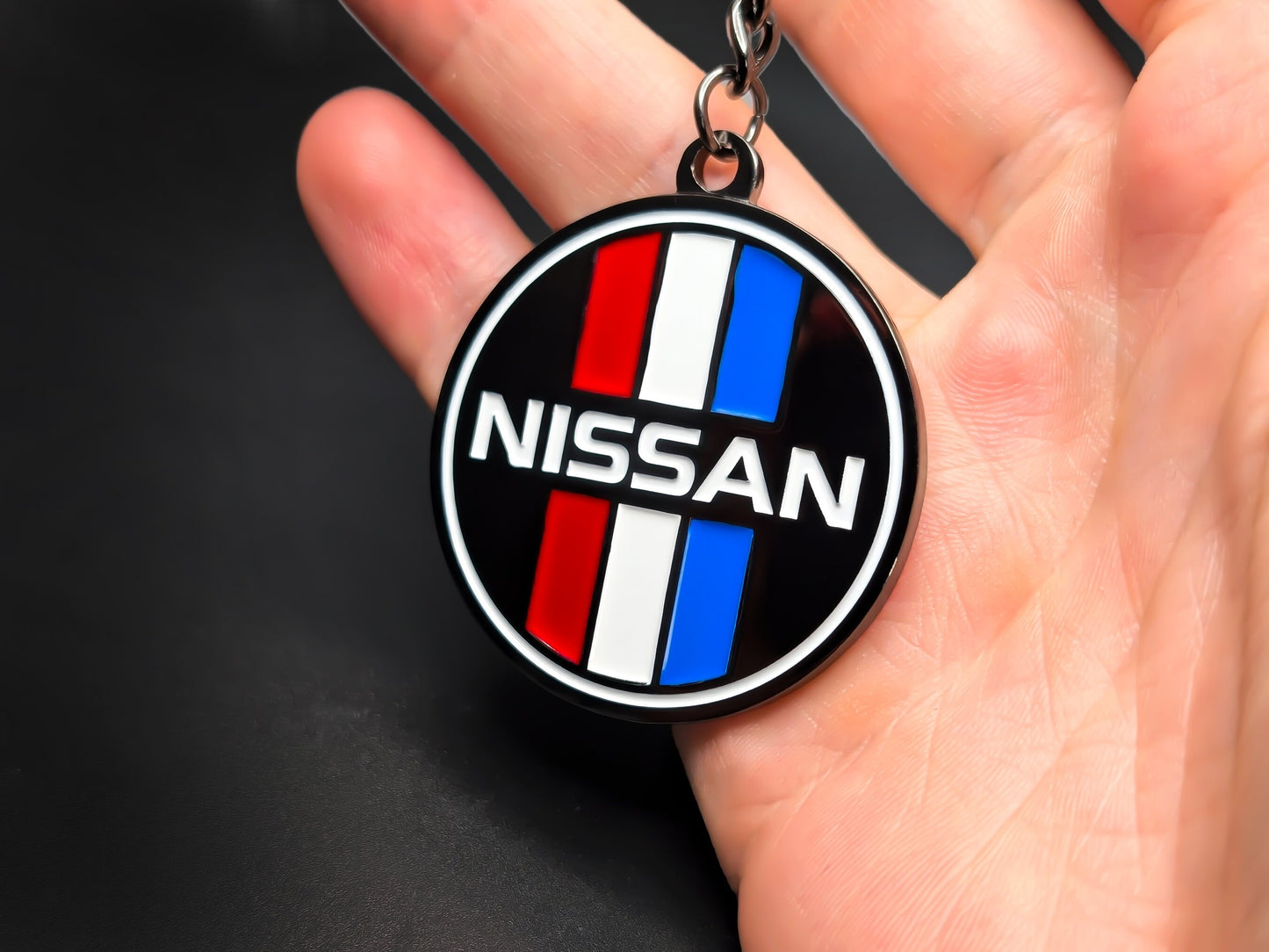 Circular Nissan heritage badge keychain held in hand, displaying the high-contrast racing colors and premium 3D raised enamel finish.