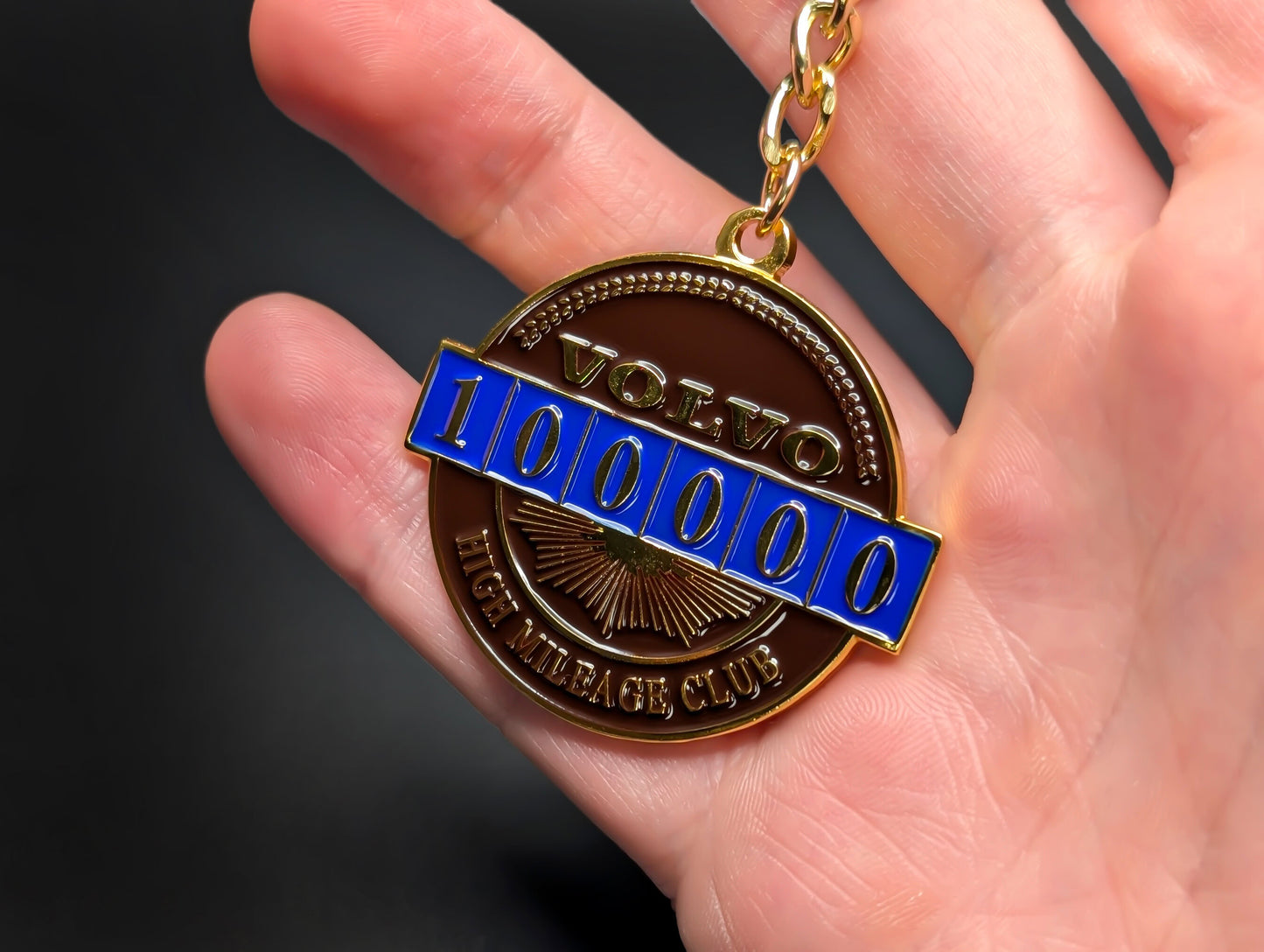 A hand holding the Volvo High Mileage Club badge keychain, showcasing the reflective gold finish and detailed 100,000 mile marker.