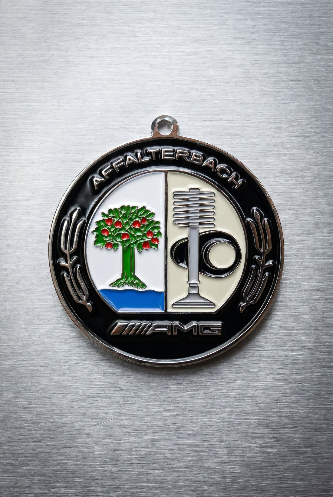 Round Mercedes-Benz AMG Affalterbach badge top-down on brushed aluminum, black border with "AFFALTERBACH" arched at top and "AMG" at bottom.