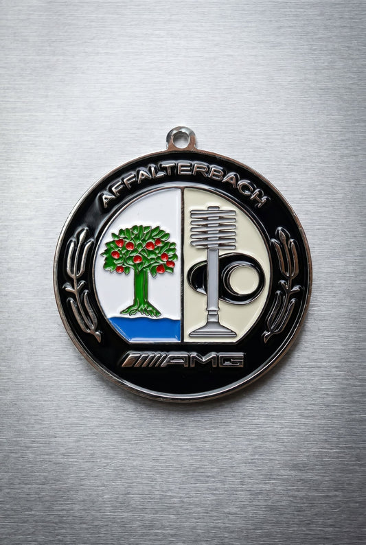 Round Mercedes-Benz AMG Affalterbach badge top-down on brushed aluminum, black border with "AFFALTERBACH" arched at top and "AMG" at bottom.