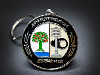 Round black and silver AMG double-sided metal keychain with the Affalterbach emblem, featuring a tree, engine part, and the AMG logo.