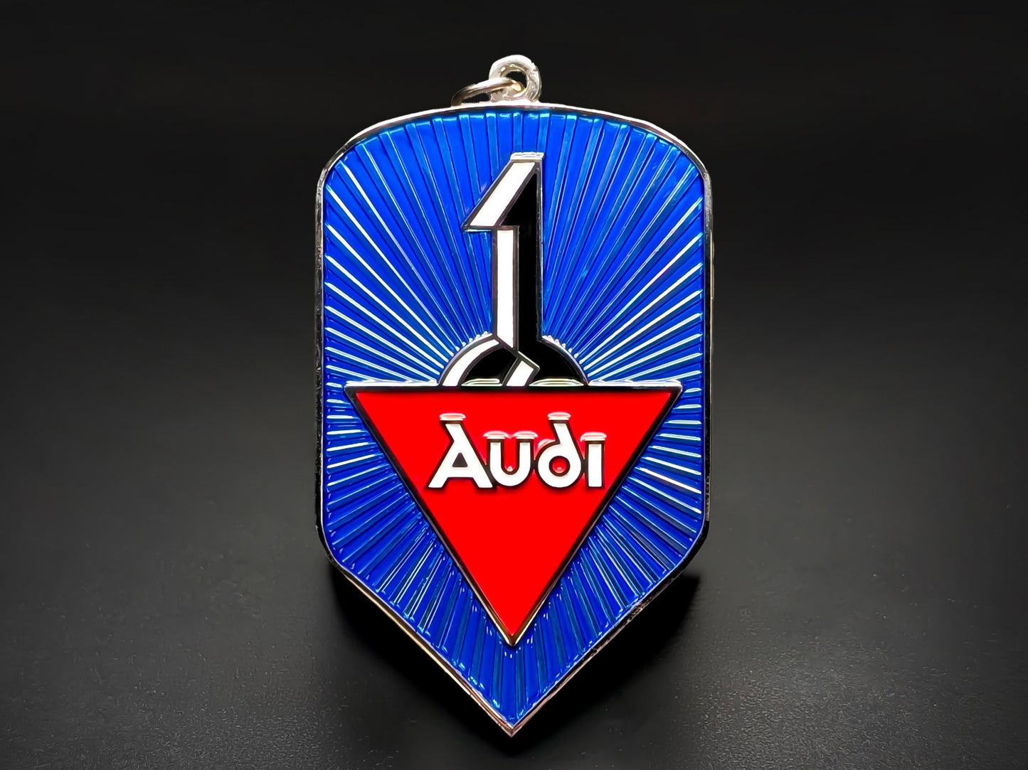 A shield-shaped metal keychain badge featuring a vibrant blue radial sunburst pattern. In the center, a red triangle contains the white stylized "Audi" script.