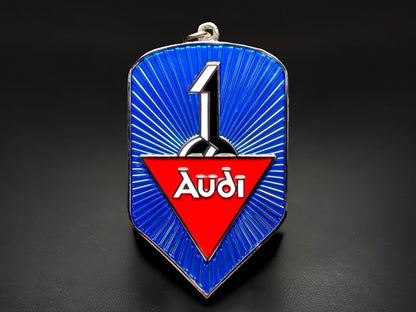 A shield-shaped metal keychain badge featuring a vibrant blue radial sunburst pattern. In the center, a red triangle contains the white stylized "Audi" script.