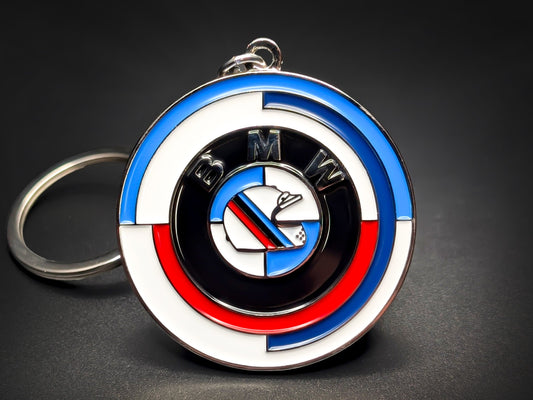 A circular metal keychain featuring the BMW name surrounded by concentric rings in the iconic Motorsport colors of light blue, dark blue, and red. The center of the design showcases a stylized white racing helmet with diagonal M-series stripes.