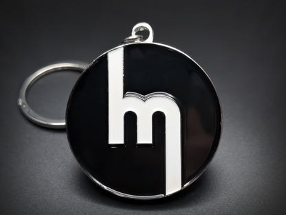 Round black double-sided metal Mazda keychain with a stylized white "M" logo.