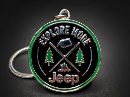 A circular, black keychain with a green rim, featuring a camping and exploration theme. It displays "EXPLORE MORE" at the top, two crossed walking sticks, a campfire, two pine trees, a tent, and the word "Jeep" at the bottom, all in silver and green.
