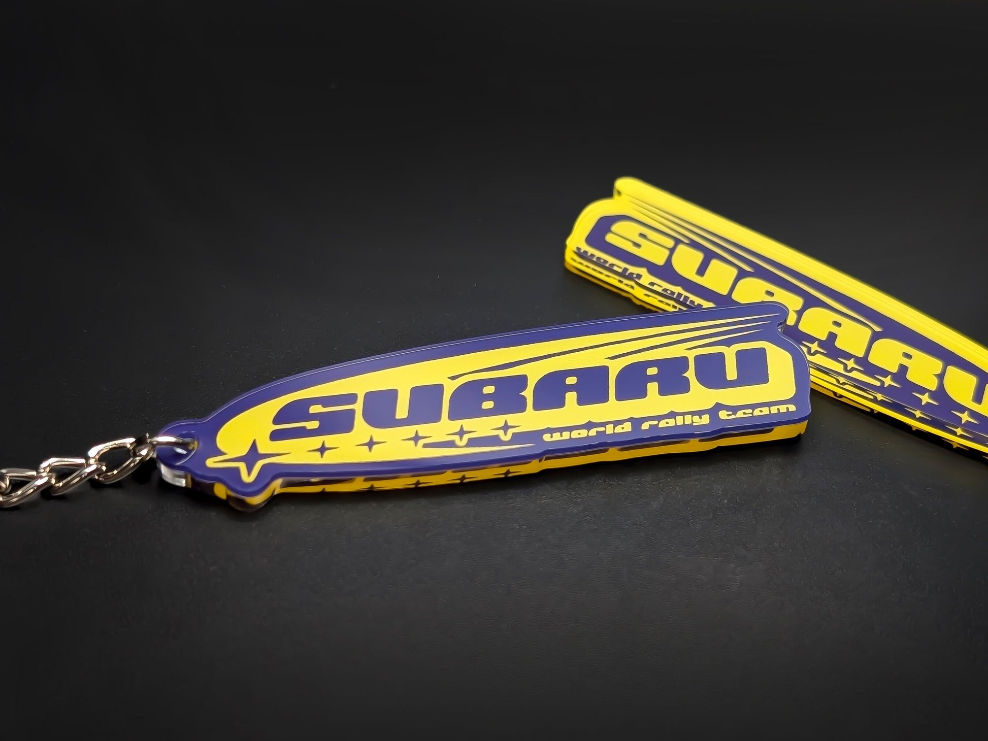 Retro blue and yellow Subaru World Rally Team acrylic keychain with "SUBARU" text and star pattern.