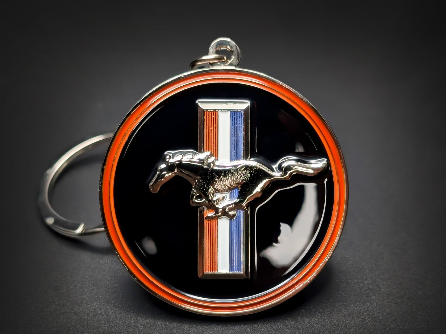 Round black and orange Ford Mustang double-sided metal keychain with a chrome horse logo and red, white, and blue racing stripes.