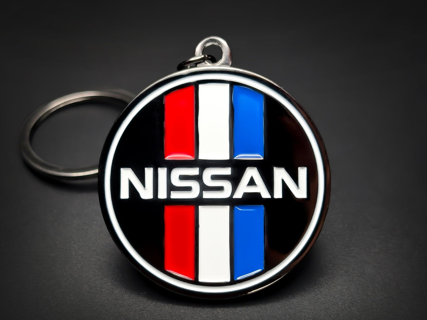 Round black retro metal Nissan keychain with red, white, and blue vertical stripes and "NISSAN" text.