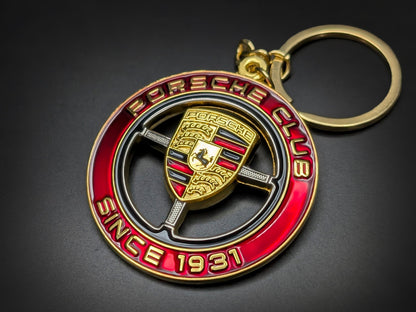 A circular metal keychain with a polished gold-tone finish and a vibrant red outer border. The red ring features the words "PORSCHE CLUB" at the top and "SINCE 1931" at the bottom in embossed gold lettering.

In the center, a gold-tone three-spoke steering wheel frame holds a detailed Porsche crest.