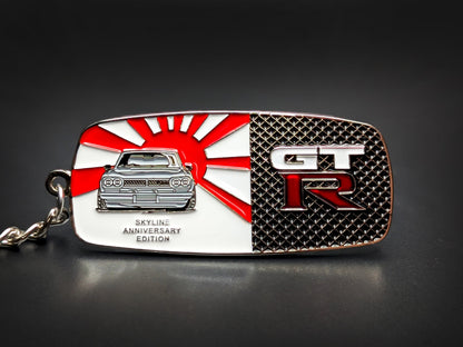 A rectangular metal keychain badge with a split design featuring a classic Nissan Skyline front-end against a red and white Rising Sun flag on the left, and a silver "GT-R" emblem over a black honeycomb mesh pattern on the right.

The text "Skyline Anniversary Edition" is embossed in silver at the bottom left.