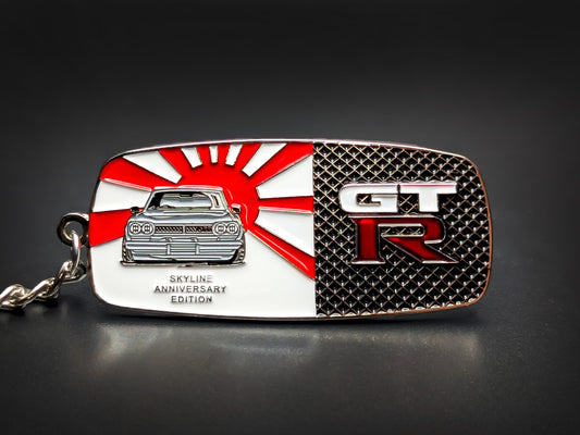 A rectangular metal keychain badge with a split design featuring a classic Nissan Skyline front-end against a red and white Rising Sun flag on the left, and a silver "GT-R" emblem over a black honeycomb mesh pattern on the right.

The text "Skyline Anniversary Edition" is embossed in silver at the bottom left.