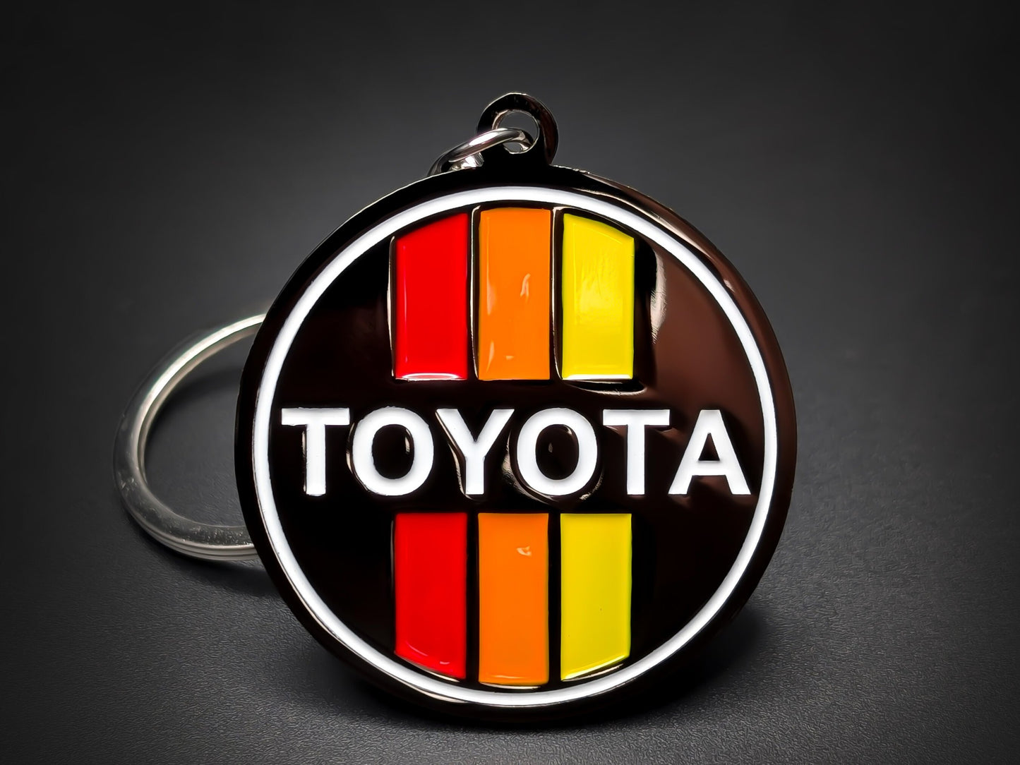 Round black metal Toyota double-sided keychain with a white logo and red, orange, and yellow vertical retro stripes.
