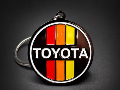 Round black metal Toyota double-sided keychain with a white logo and red, orange, and yellow vertical retro stripes.
