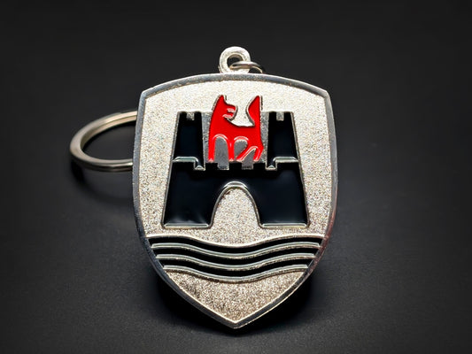 A shield-shaped metal keychain with a textured silver-glitter background. It features an embossed black castle gateway with three towers, and a vibrant red wolf standing on the central battlement.

The bottom of the shield is accented with three black horizontal wavy lines, and the piece is attached to a polished silver-tone metal ring.
