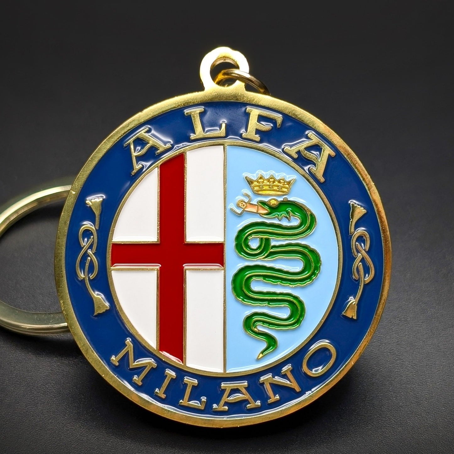 Double-sided metal Alfa Romeo badge keychain, a gold roundel bordered in blue with ‘ALFA’ and ‘MILANO’ text, a red cross and green serpent crest at center, on a silver split ring on a dark surface.