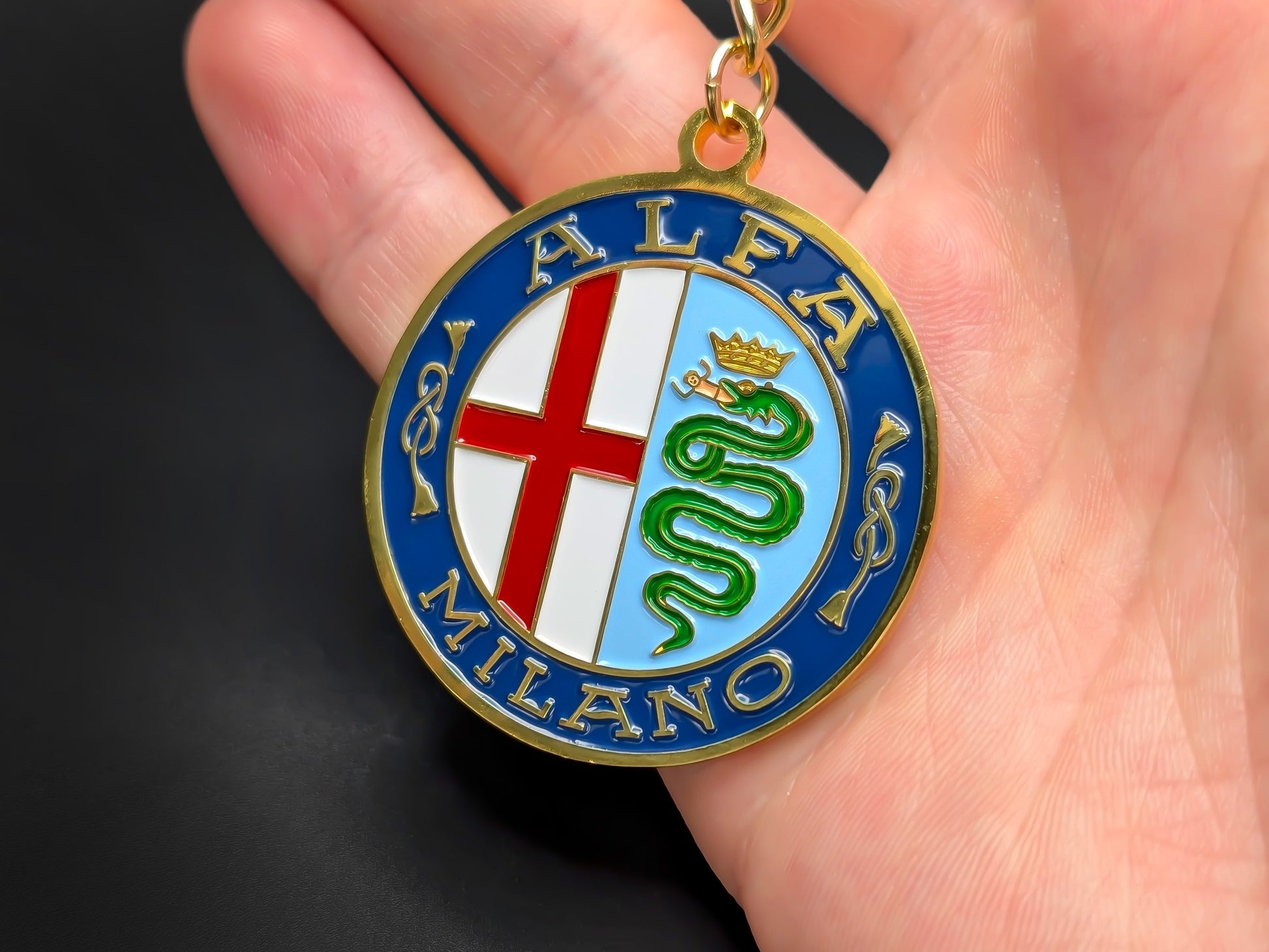 Hand holding a circular Alfa Romeo Milano enamel badge keychain to show the premium weight and 3D raised gold-plated details.