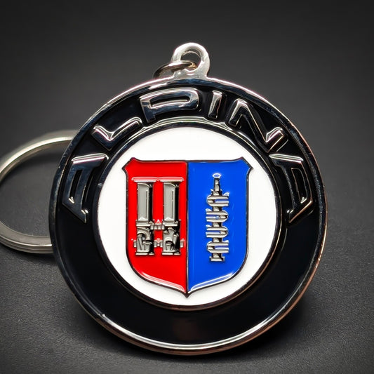 Double-sided metal Alpina badge keychain with a black ring reading ‘ALPINA’ in silver, circling a white shield split red and blue with silver line-art emblems, chrome split ring, dark background.