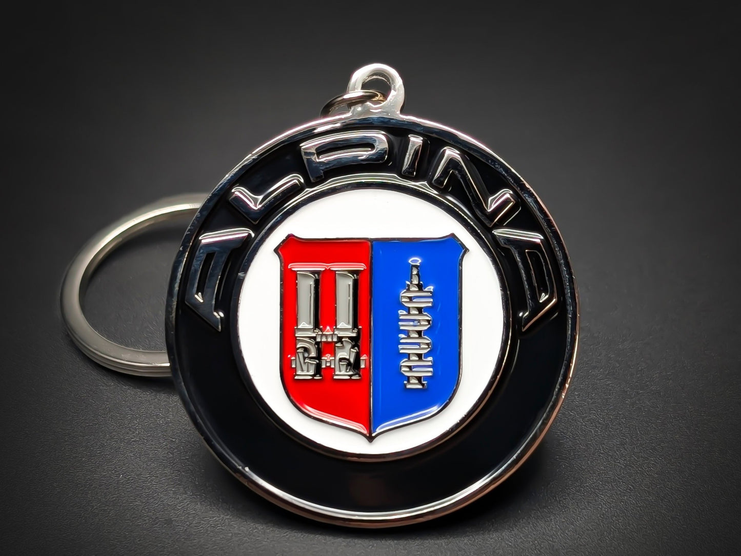 Vintage Alpina badge keychain featuring the red and blue heritage shield with silver crankshaft and carburetor details on a black border.