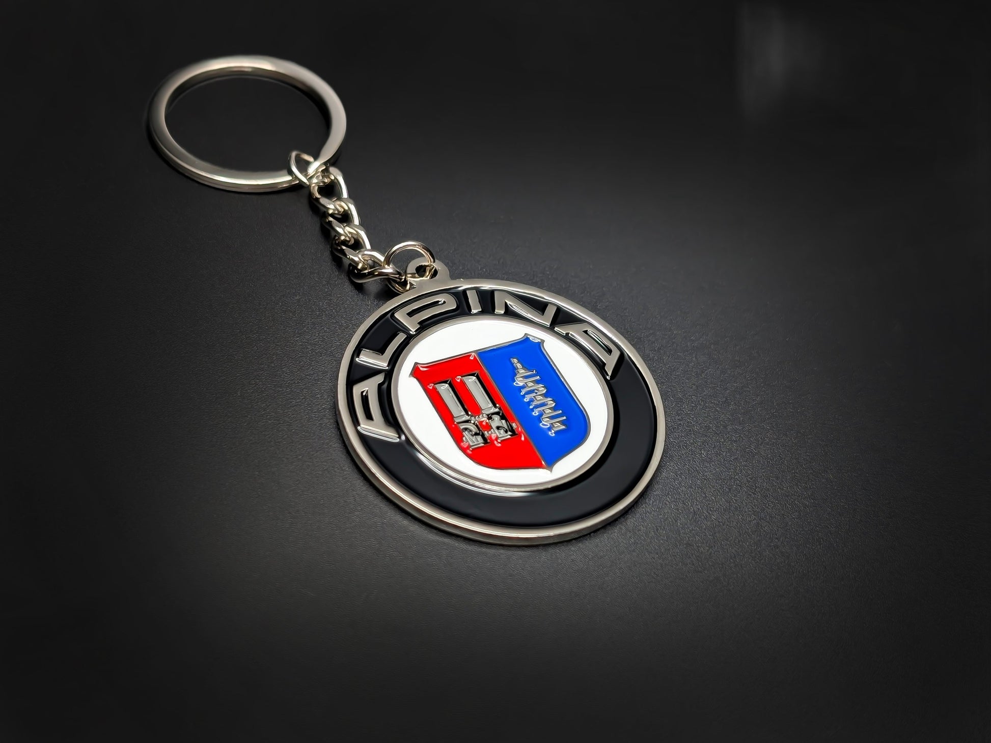 High-quality Alpina heritage badge keychain on a dark background, showcasing the polished silver-tone lettering and glossy enamel finish.