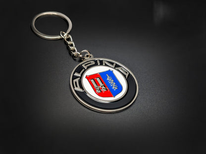 High-quality Alpina heritage badge keychain on a dark background, showcasing the polished silver-tone lettering and glossy enamel finish.