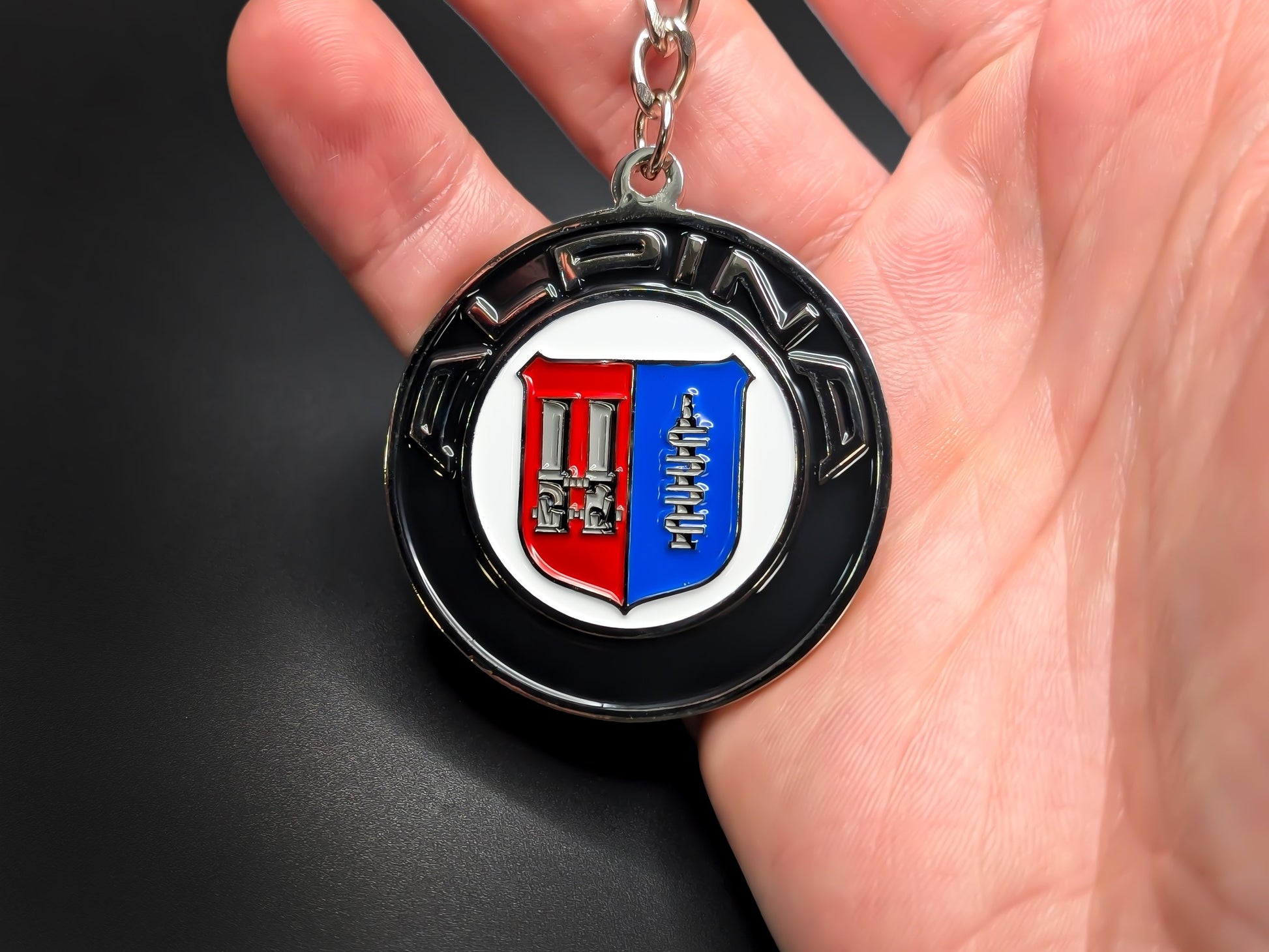Hand holding a circular Alpina enamel badge keychain to demonstrate the premium metallic weight and 3D raised silver details.