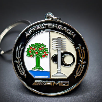 Double-sided metal Mercedes-AMG badge keychain, black and chrome medallion reading “Affalterbach” at top and “AMG” at bottom, a tree-and-water crest and tool-and-wheel design, silver split ring.