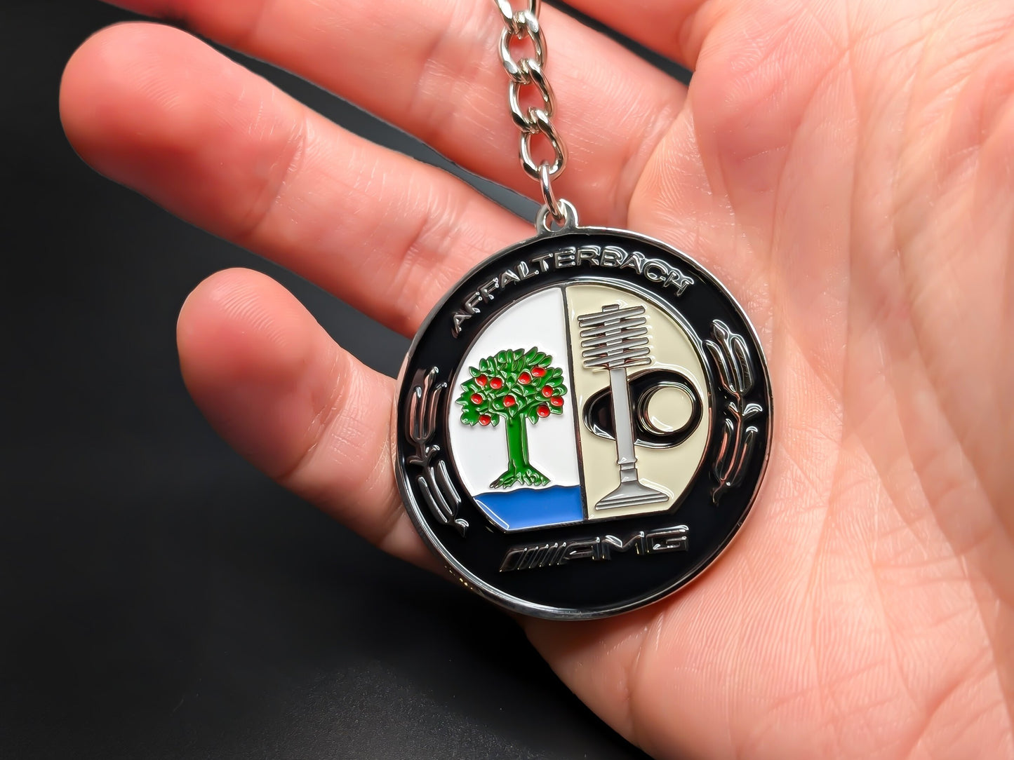 Hand holding a circular AMG Affalterbach enamel badge keychain to demonstrate the premium metallic weight and detailed 3D color finish.