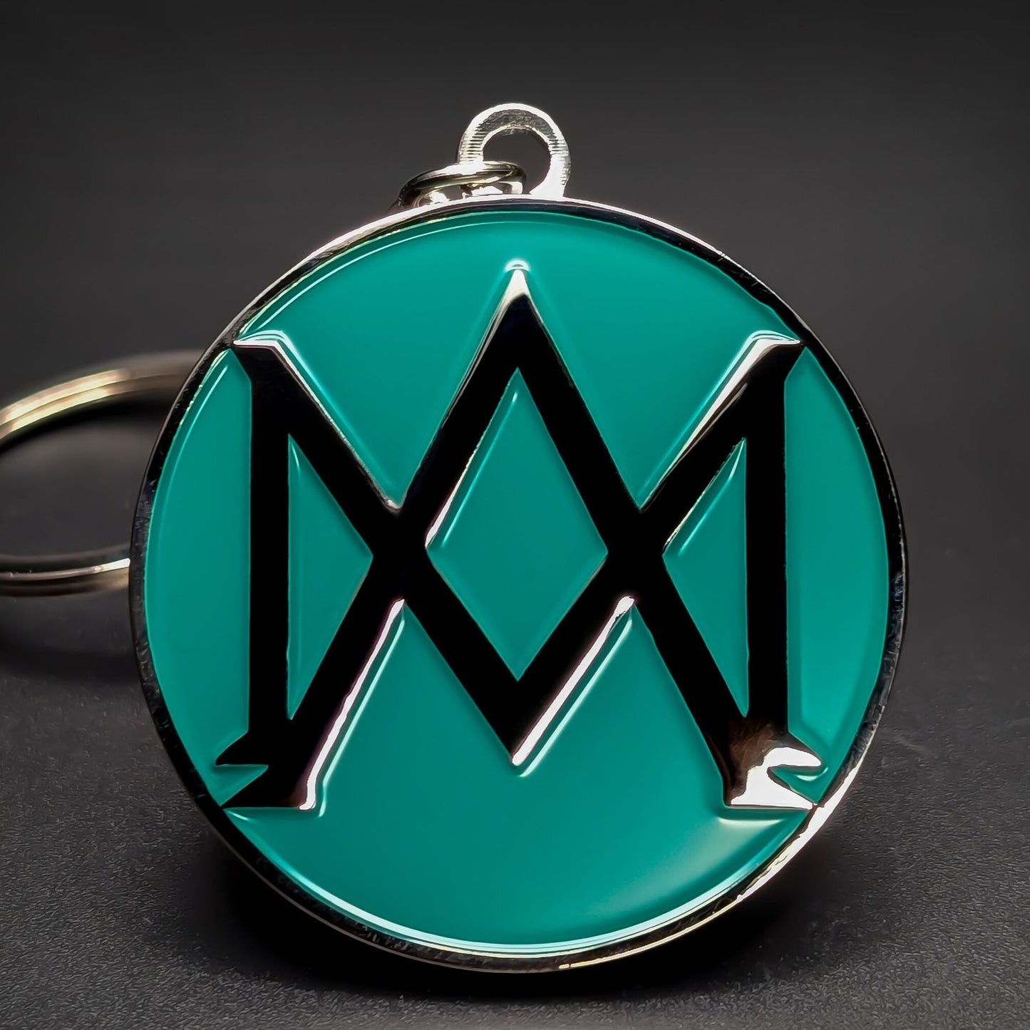 Double-sided metal Aston Martin badge keychain with a black abstract wing emblem on a glossy green enamel background, chrome rim and loop at the top, on a dark gradient surface.