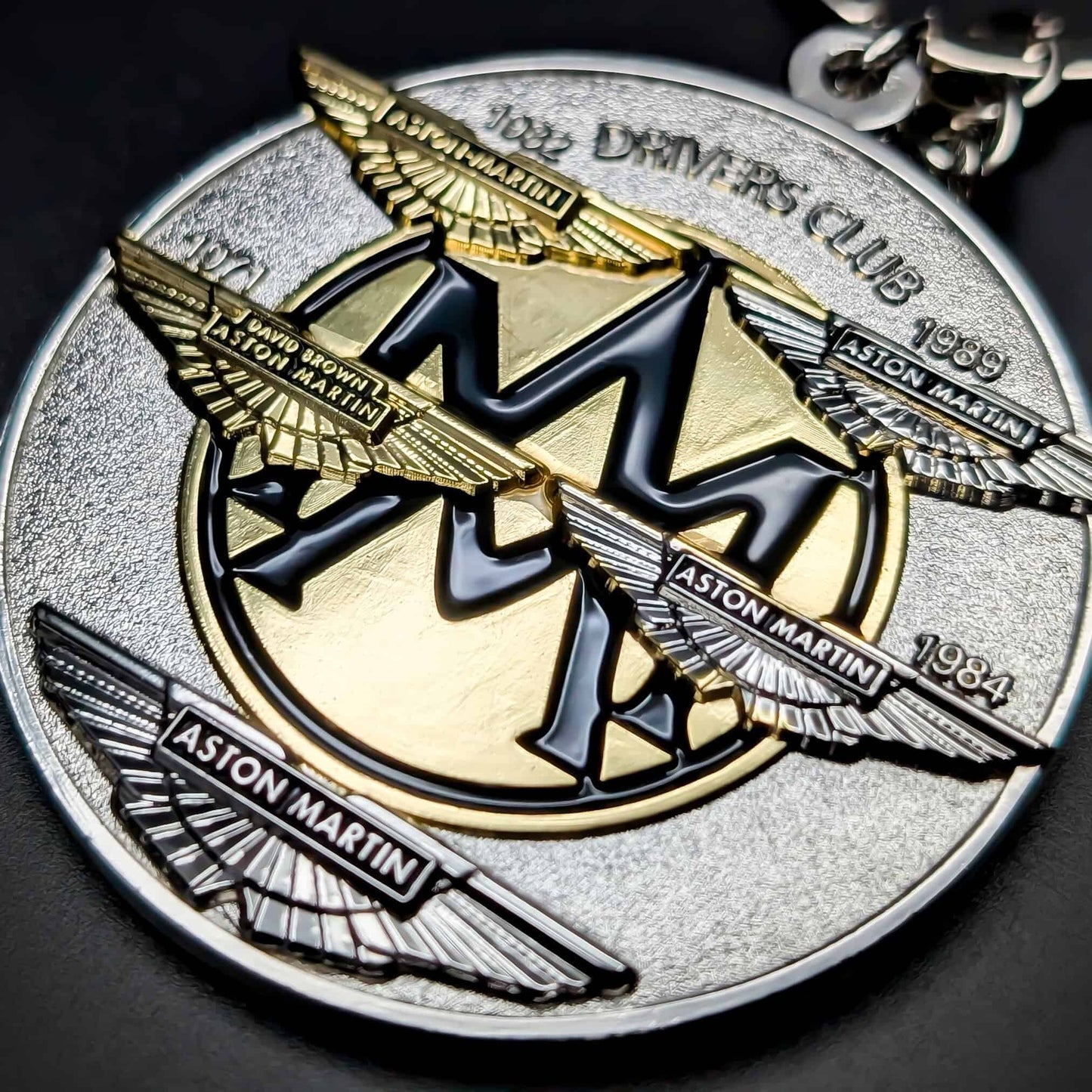 Double-sided round metal Aston Martin badge keychain, textured silver face with layered gold, gunmetal, and silver winged Aston Martin logos, ASTON MARTIN nameplates, chrome chain at the edge.