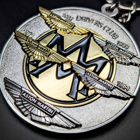 Double-sided round metal Aston Martin badge keychain, textured silver face with layered gold, gunmetal, and silver winged Aston Martin logos, ASTON MARTIN nameplates, chrome chain at the edge.