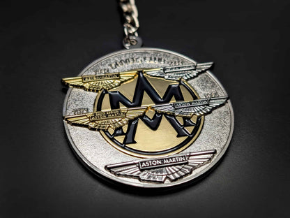 Perspective shot of the circular Aston Martin heritage badge keychain, highlighting the 3D raised metal wings and textured base.