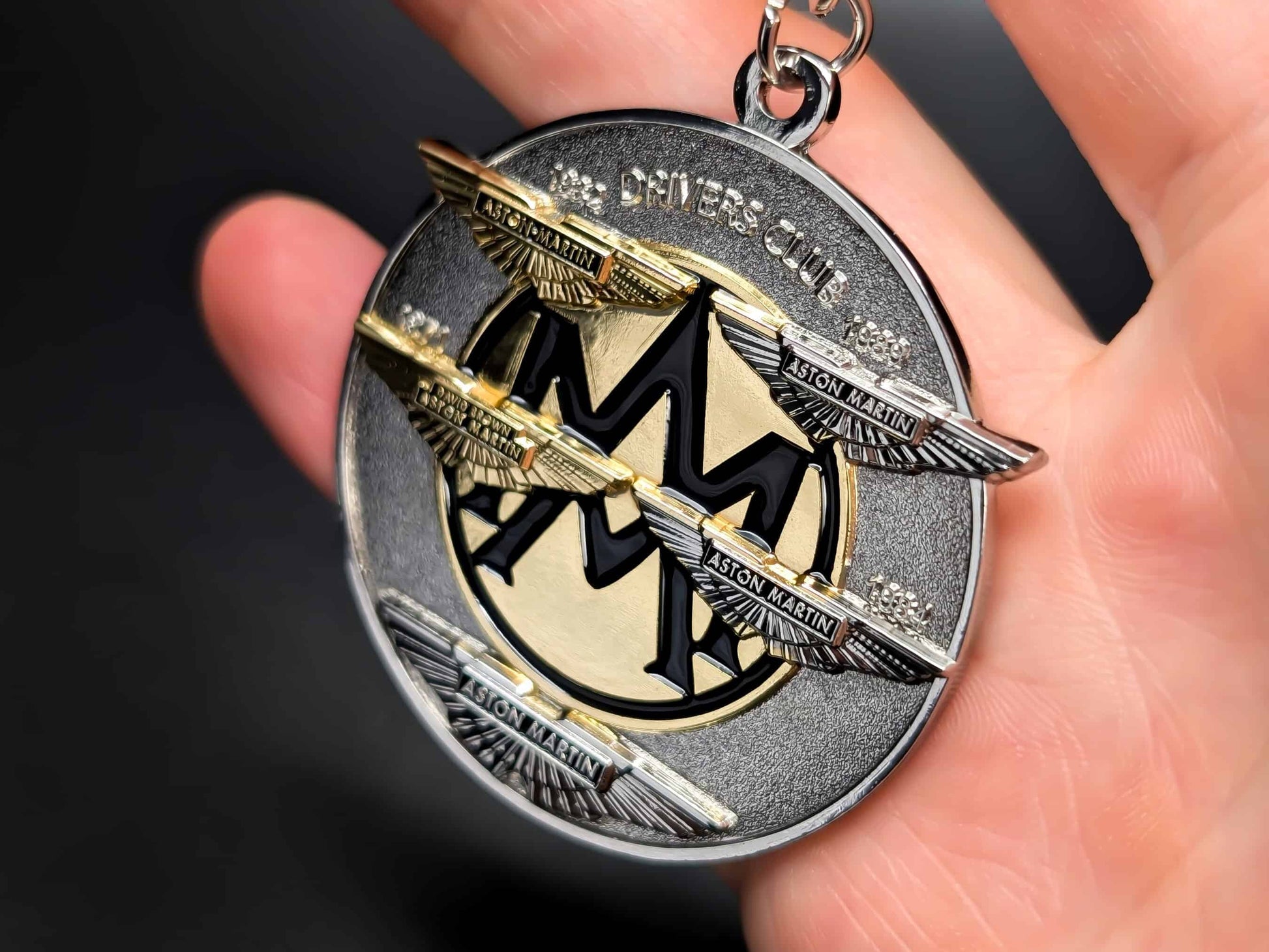 Aston Martin Drivers Club metal badge keychain held in hand to show size, featuring the iconic wings and "Drivers Club" text.