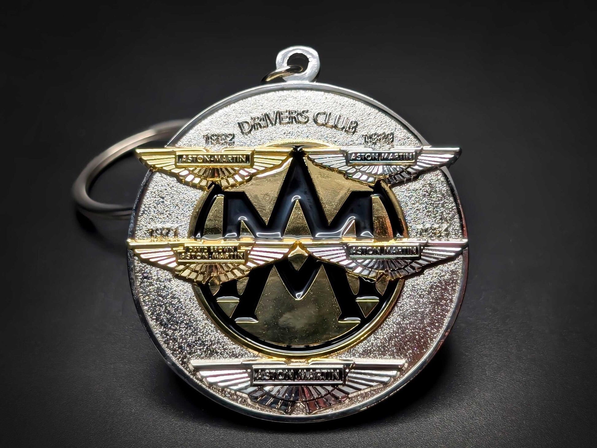 Premium silver and gold Aston Martin car badge keychain with a clean display of the central crest and anniversary dates.