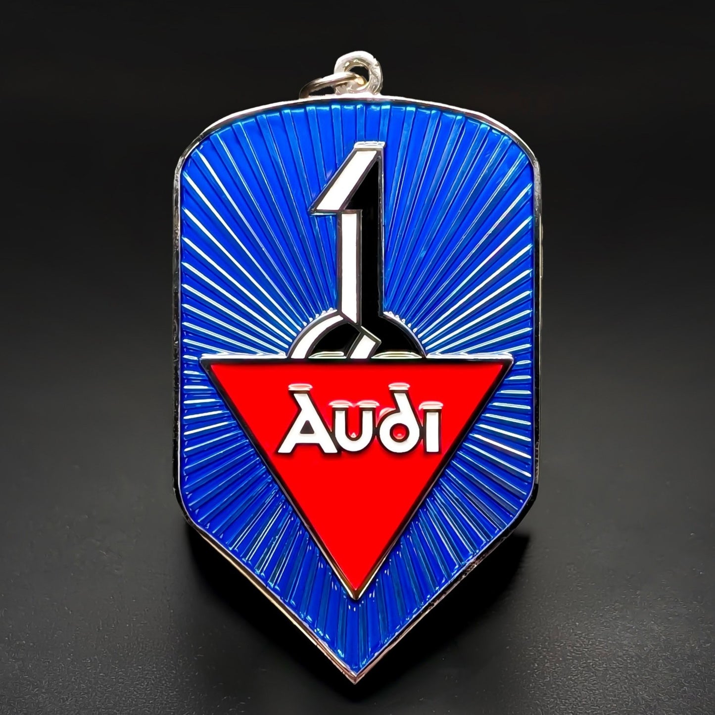 Double-sided shield-shaped metal Audi badge keychain, blue face with radiating chrome sunburst lines, a silver number 1 emblem, and a red triangle with white Audi script, chrome border and hanging loop.