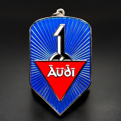 Double-sided shield-shaped metal Audi badge keychain, blue face with radiating chrome sunburst lines, a silver number 1 emblem, and a red triangle with white Audi script, chrome border and hanging loop.