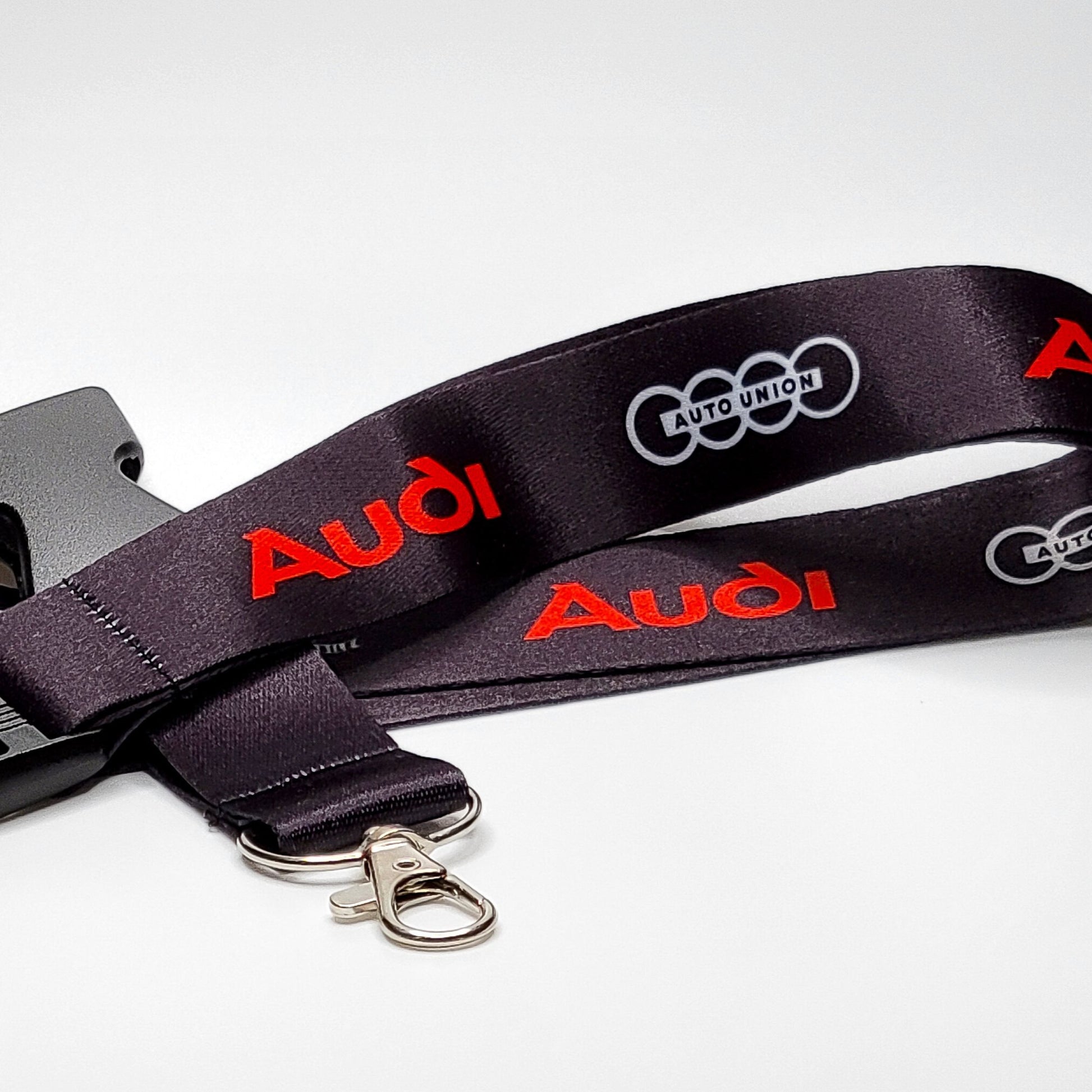 Black Audi lanyard with a red Audi logo and a grey four-ring Auto Union emblem repeated along the strap, coiled with a grey breakaway clip, silver clasp, and key ring on white.