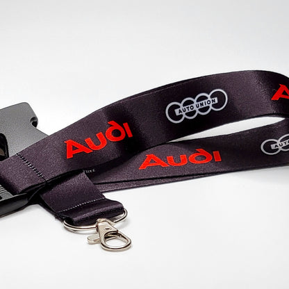 Black Audi lanyard with a red Audi logo and a grey four-ring Auto Union emblem repeated along the strap, coiled with a grey breakaway clip, silver clasp, and key ring on white.