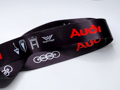Close-up of a black satin lanyard showing historical Audi brand logos including Horch, Wanderer, and DKW.