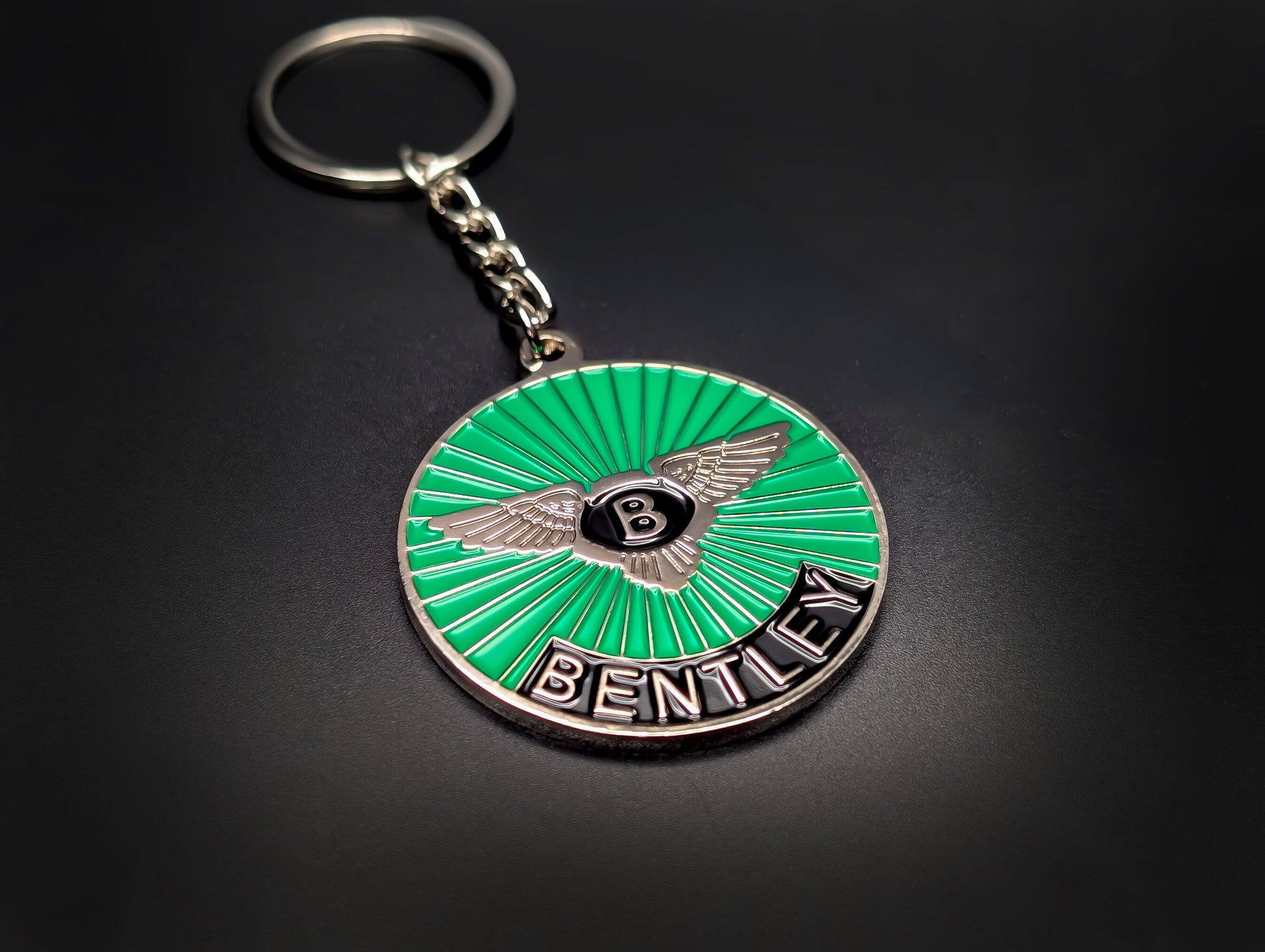 A green Bentley sunburst badge keychain with a silver chain and ring, showcased against a smooth dark background.