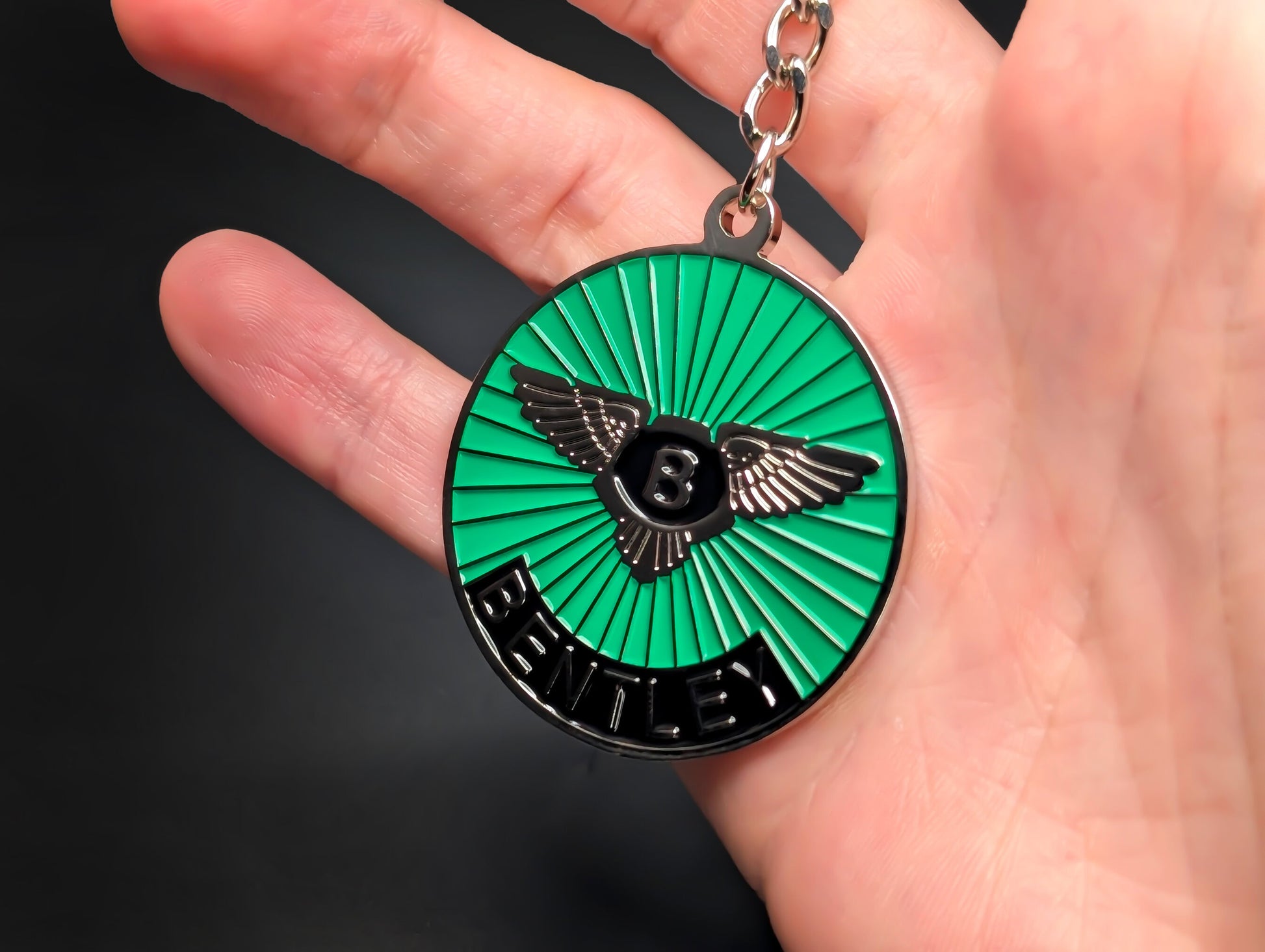 A hand holding the green Bentley badge keychain to demonstrate the scale and the detailed black and silver "Bentley" lettering.