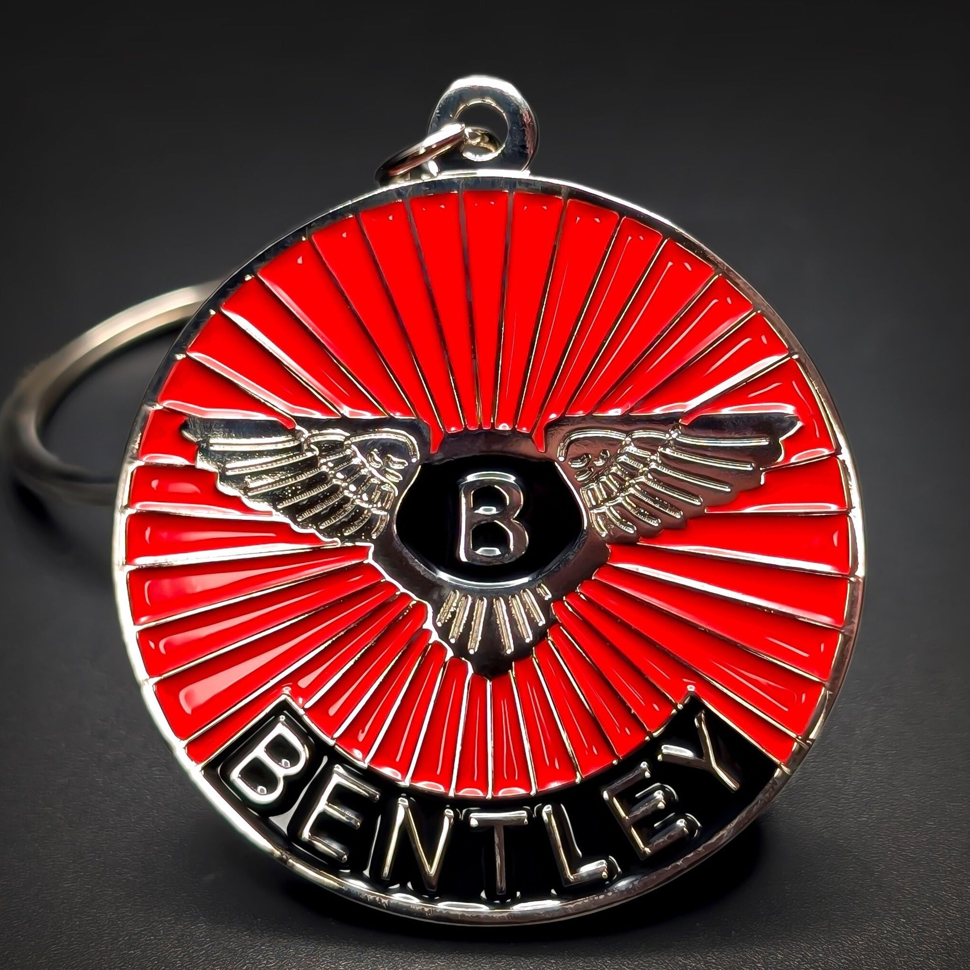 Double-sided metal Bentley badge keychain with silver wings and a silver B on a black shield over red sunburst enamel, BENTLEY lettering on a black band, silver split ring, on a dark surface.