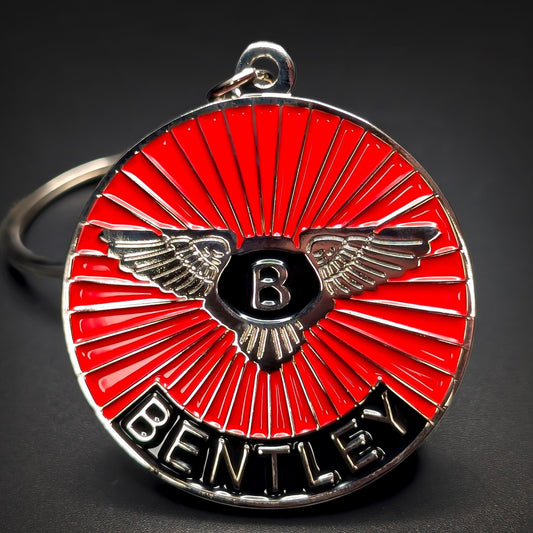 Double-sided metal Bentley badge keychain with silver wings and a silver B on a black shield over red sunburst enamel, BENTLEY lettering on a black band, silver split ring, on a dark surface.