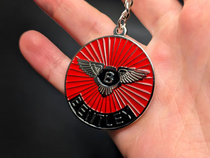 A hand holding the Bentley badge keychain, highlighting the polished metal finish and the detailed "Bentley" lettering at the bottom.