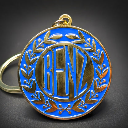 Double-sided metal Benz badge keychain, round gold-tone medallion with a blue enamel field, a gold laurel wreath border, and “BENZ” lettering at center, gold-tone split ring.