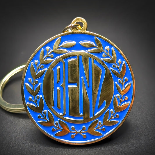 Double-sided metal Benz badge keychain, round gold-tone medallion with a blue enamel field, a gold laurel wreath border, and “BENZ” lettering at center, gold-tone split ring.